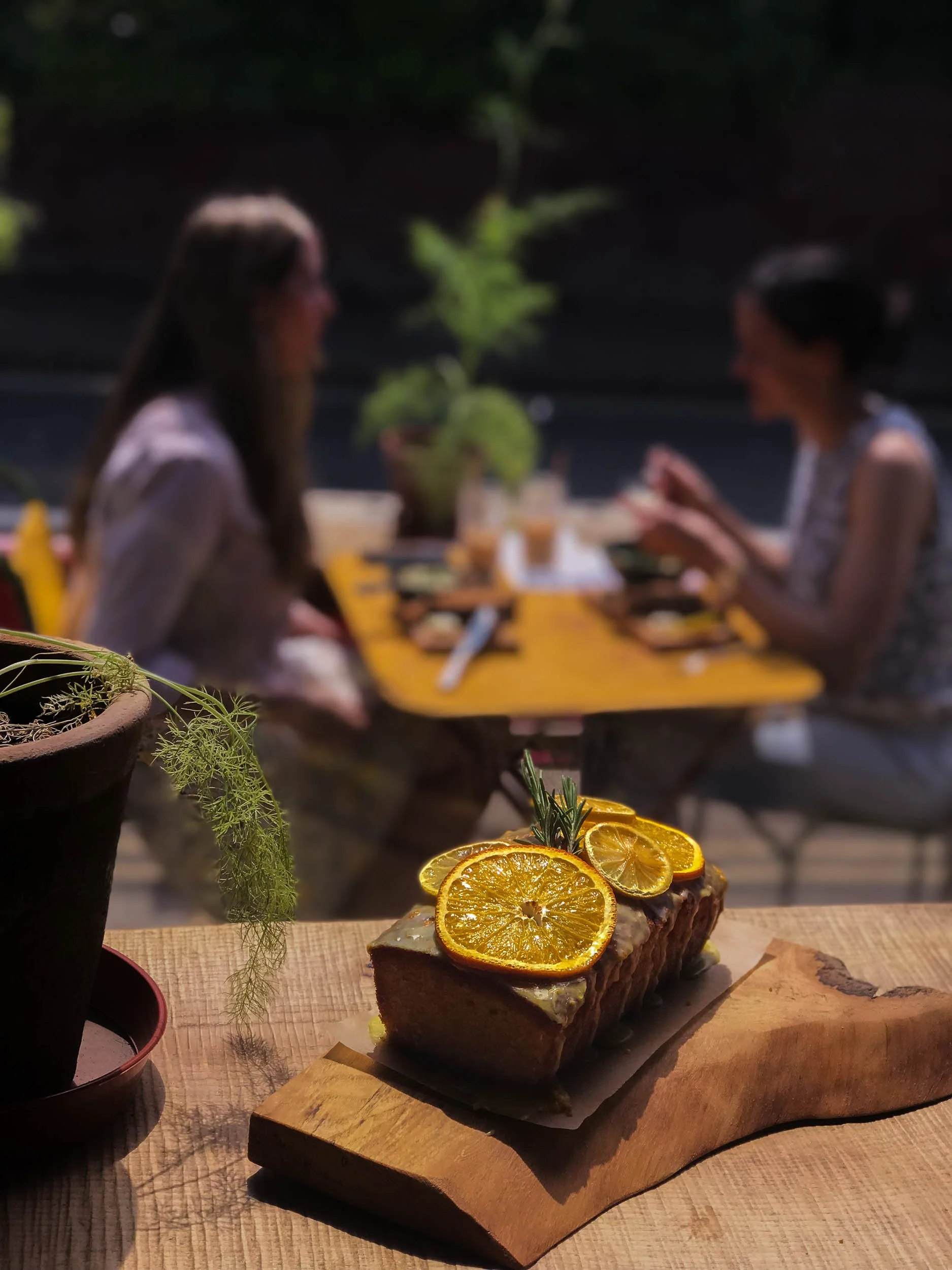A slice of cake topped with dried orange slices and herbs, placed on a wooden serving board. In the background, two women are sitting at a table outdoors, talking and smiling with greenery behind them.