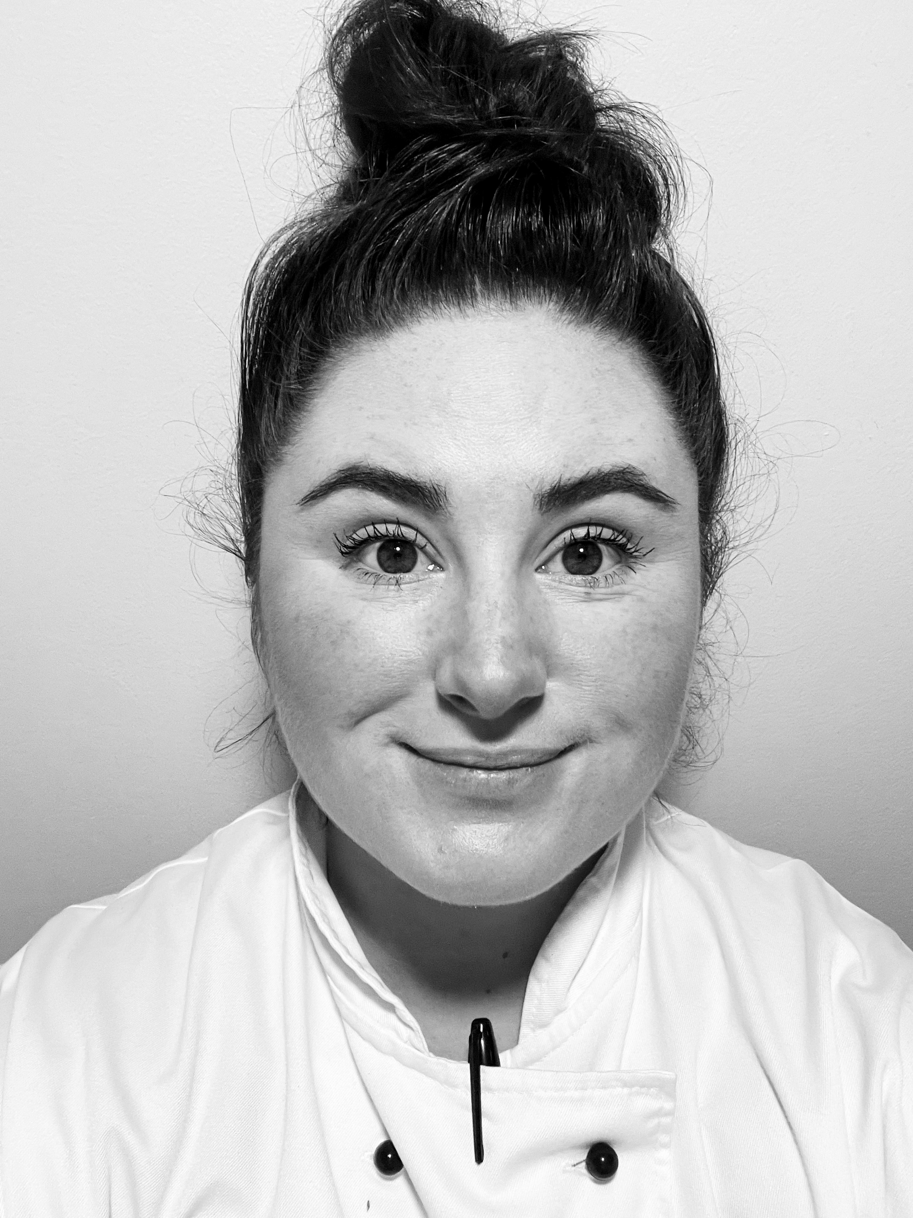 Chef Olivia Carbonari with dark hair tied up in a messy bun, smiling, wearing a chef's coat with black buttons, holding a pen, against a plain wall.