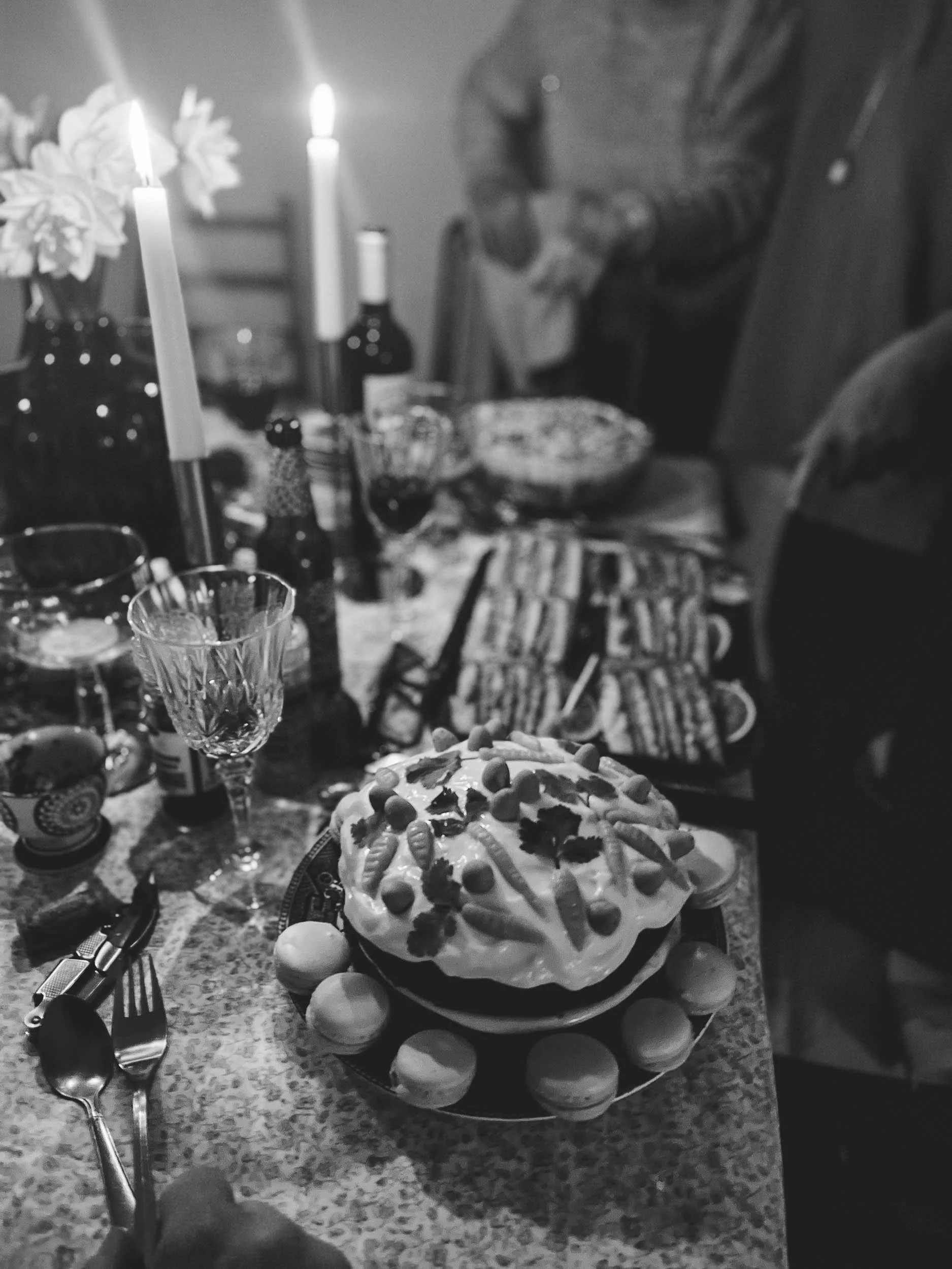 A birthday celebration scene in black and white with a birthday cake decorated with various candies and icing in the center. The table also has several empty glasses, bottles, and utensils. In the background, a person is standing with hands clasped, and there are lit candles and flowers on the table.