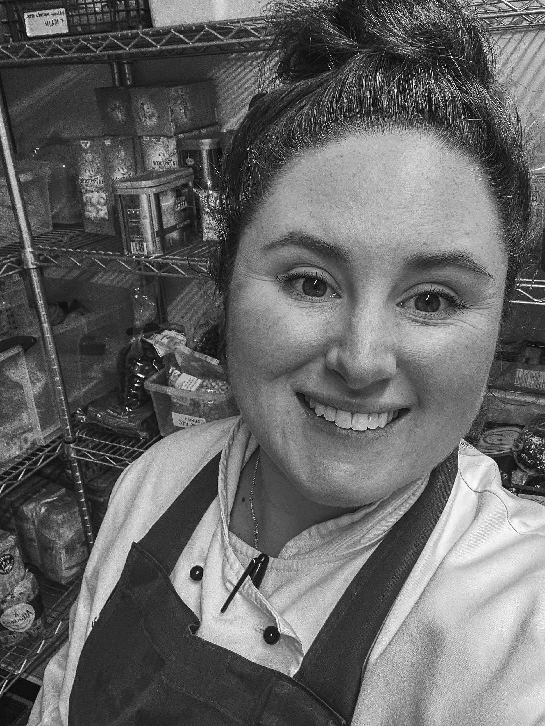 Chef Olivia Carbonari in frame with dark hair in a messy bun, smiling, wearing chef white jacket and apron standing in storage room with metal shelves filled with food items.