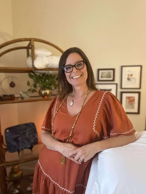 Woman with glasses and brown hair smiling, wearing a rust-colored dress with white embroidery, standing in a cozy room with framed pictures on the wall, a bookshelf, and decorative plants.