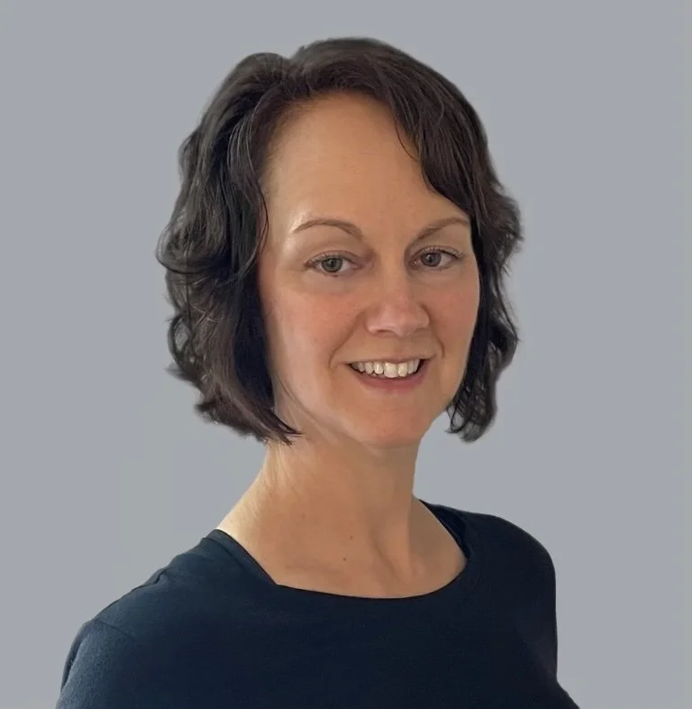 A woman with short, wavy dark hair wearing a black top, smiling, against a plain, light gray background.