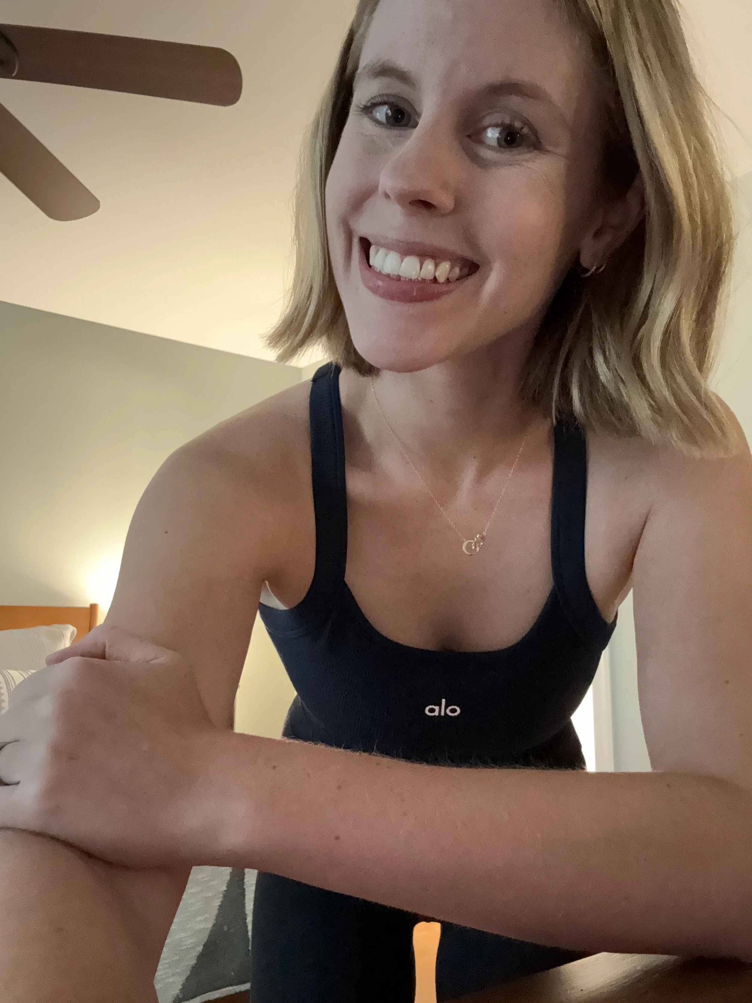 A smiling woman with shoulder-length blonde hair taking a selfie in a room with a ceiling fan and bed in the background.