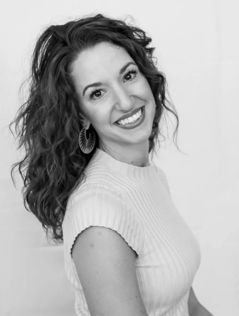 A woman with curly hair smiling and wearing a sleeveless, ribbed top and large hoop earrings, posing against a plain background.