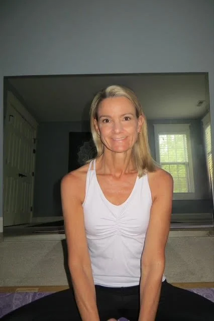 A woman with blonde hair sitting cross-legged in a room with natural light, wearing a white sleeveless top.