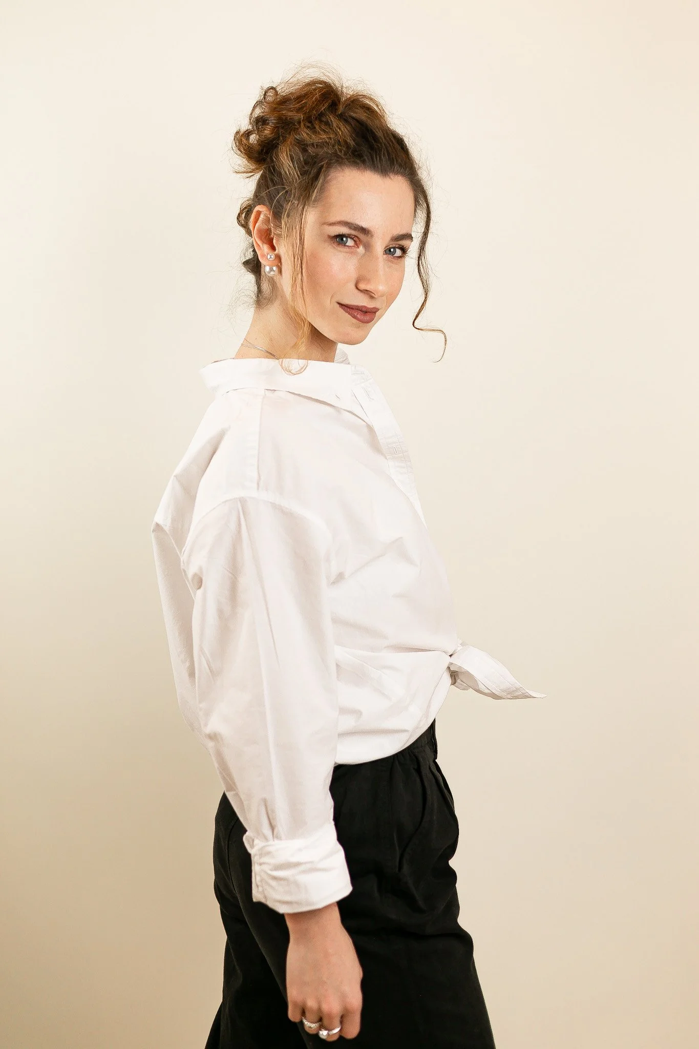 A woman with styled hair tied up in an updo, wearing a white shirt tied at the waist and black pants, standing against a plain light-colored background.