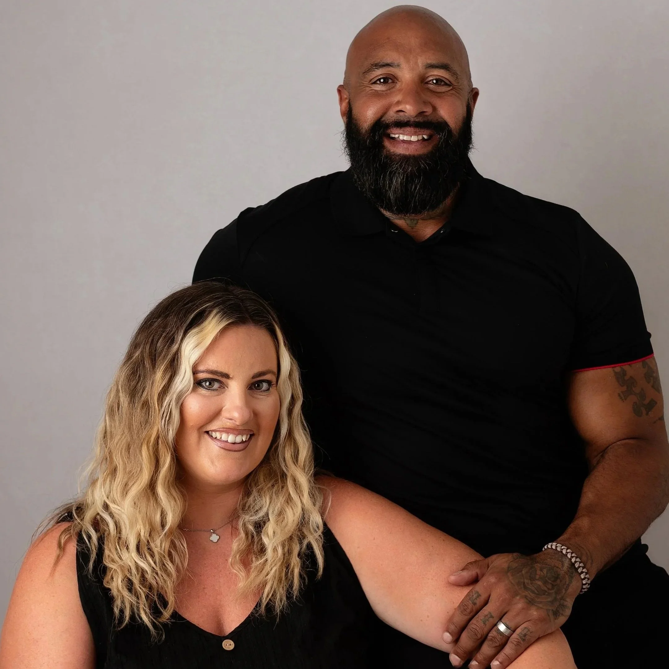 A woman with blonde, curly hair smiling as a man with a beard and bald head, wearing a black shirt, standing behind her, place his hand on her shoulder.