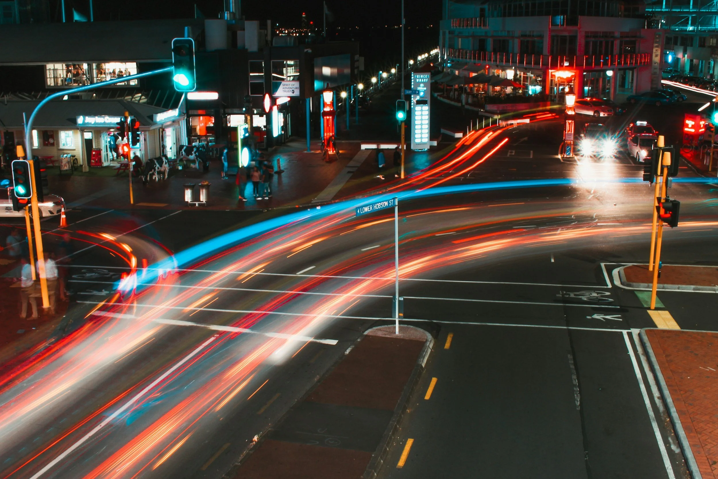 Nighttime city street with light trails from moving cars, traffic signals, pedestrians, and buildings with illuminated signs.