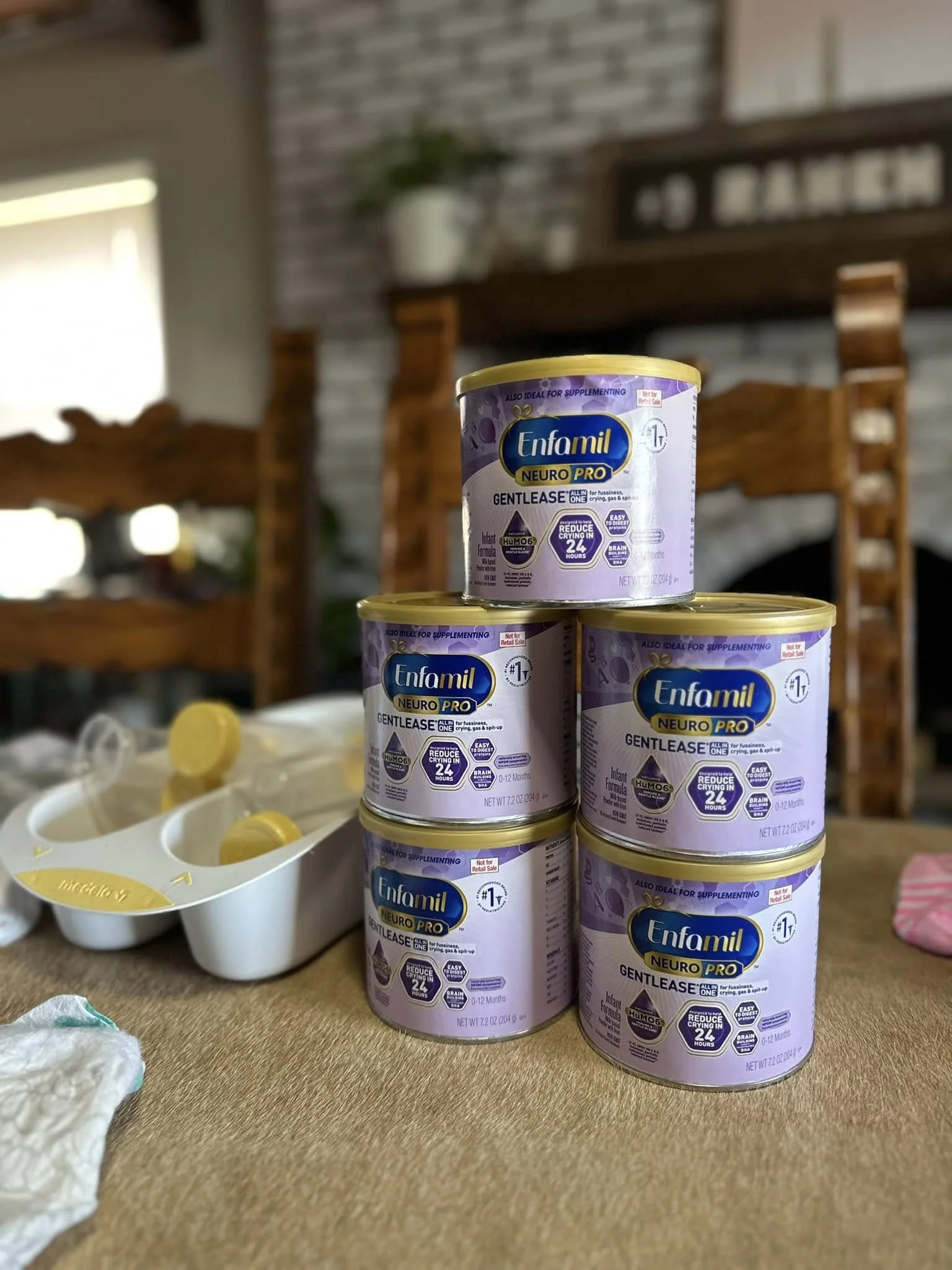 Stack of four cans of Enfamil NeuroPro Gentlease infant formula on a wooden surface in a home setting, with a white dish and some paper or cloth nearby.