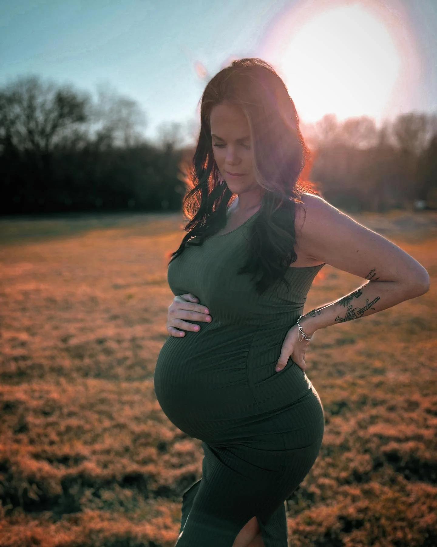 A pregnant woman standing outdoors during sunset, cradling her belly with her left hand and resting her right hand on her lower back, wearing a green sleeveless dress with tattoos on her left arm.