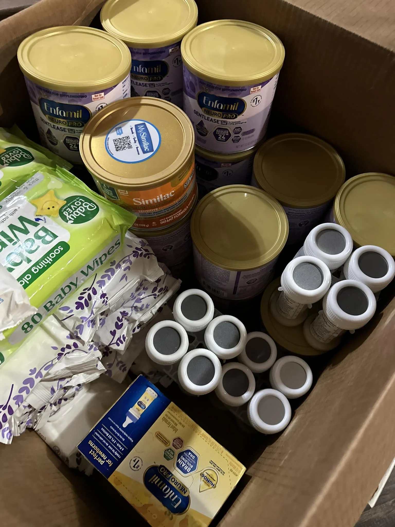 A brown paper grocery box containing several cans of Enfamil NeuroPro Infant Formula, a pack of Baby Wipes, and a box of Pedialyte electrolyte solution bottles.
