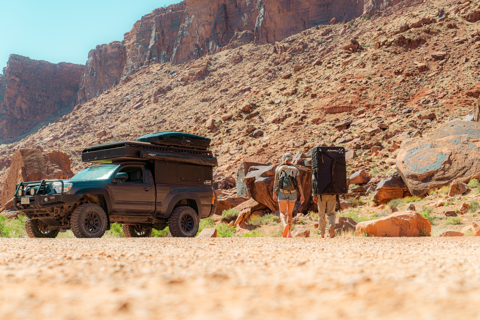 Off-road vehicle parked in rocky desert terrain with two hikers carrying backpacks, approaching a cliff.