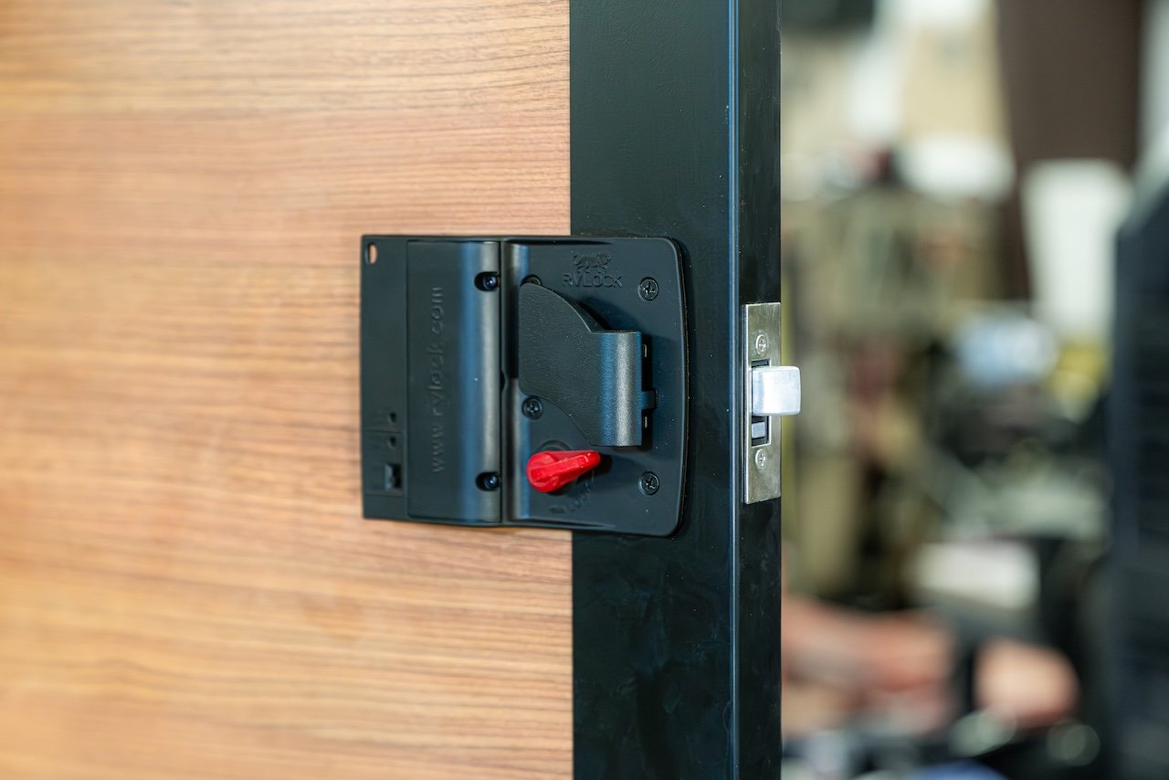 Close-up of an RV lock on a wooden door, featuring a metal latch and a black locking mechanism with a red switch.