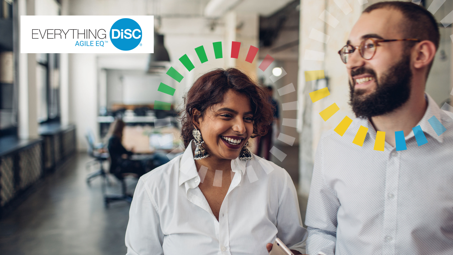 Two smiling people, a woman with short curly hair and earrings and a man with glasses and a beard, engage in conversation in a modern office setting.