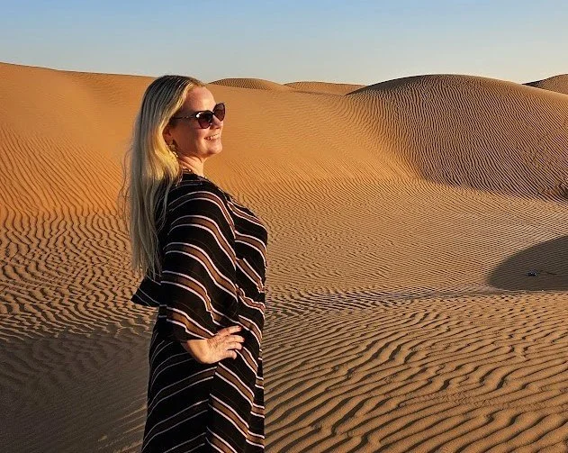 A woman with long blonde hair, sunglasses, and a striped dress standing in a desert with sand dunes.