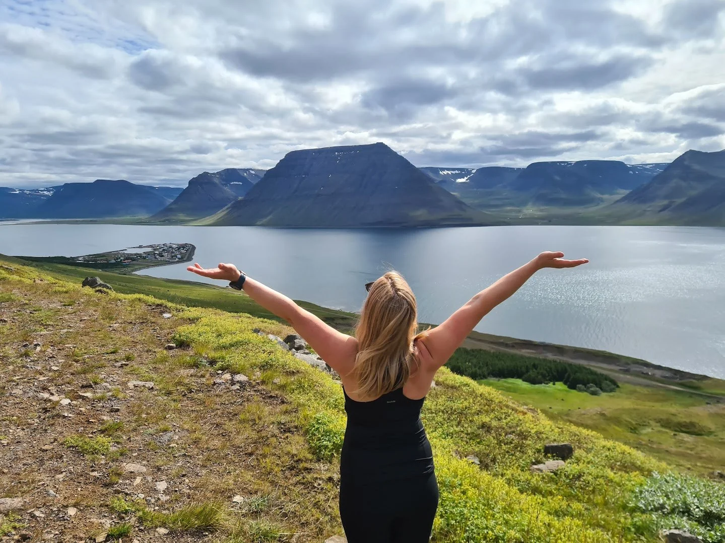 Person standing with arms raised, overlooking a fjord with mountains in the background and a small town on a narrow peninsula.