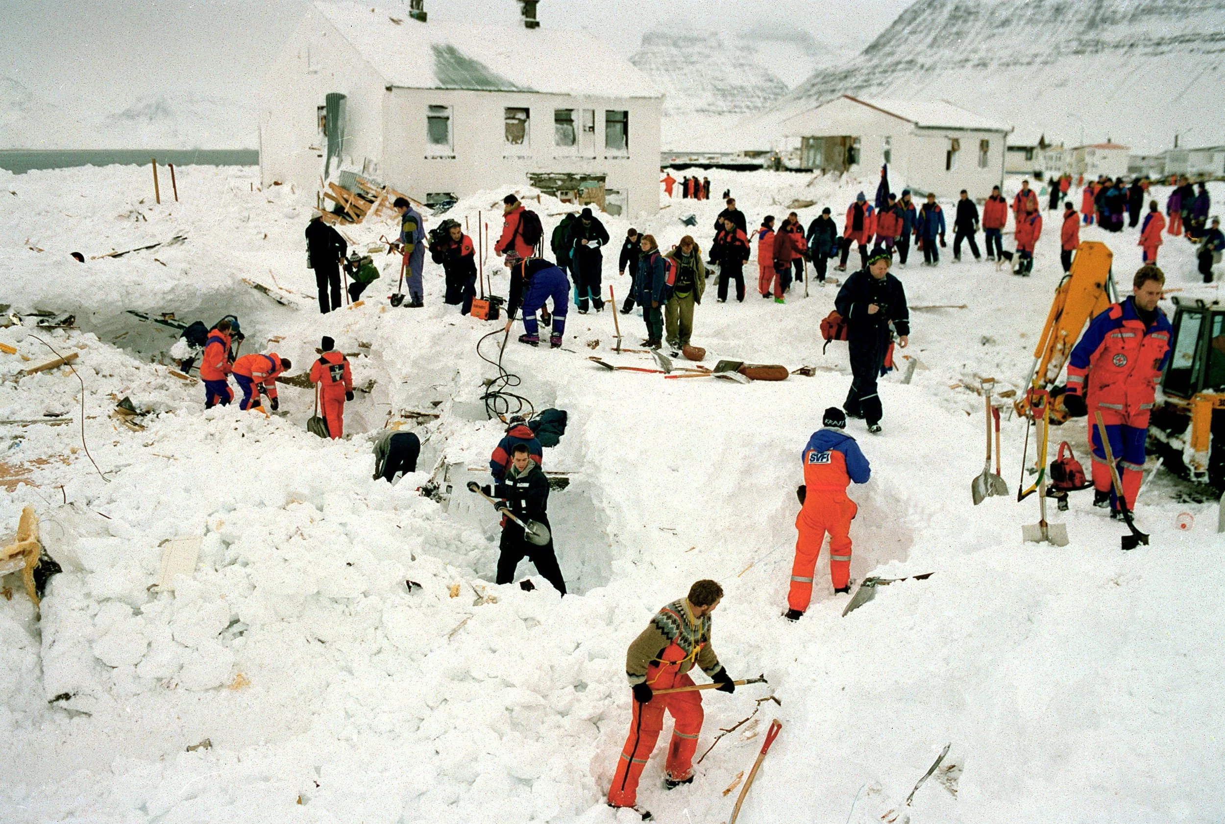 Rescue workers in red suits dig through snow after avalanche in Flateyri, mountains in background.