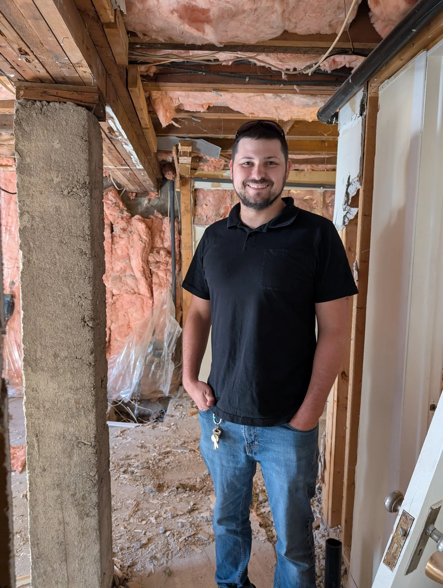 A man with a beard and short dark hair standing inside a house under renovation, smiling, wearing a black polo shirt and blue jeans, with keys hanging from his pocket.