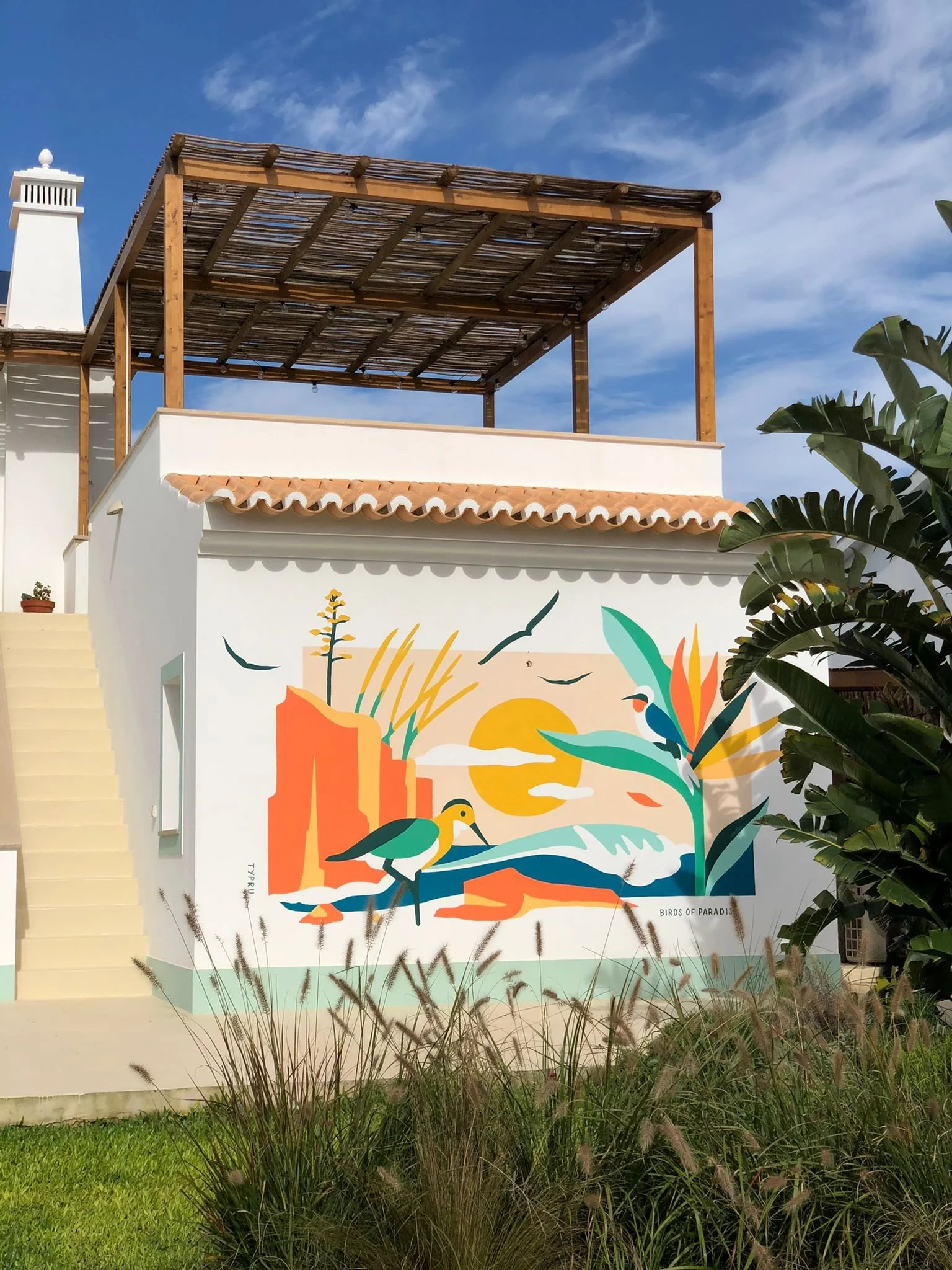 Colorful mural of a tropical landscape with birds, cliffs, mountains, and a setting sun on a building wall, with a partially open wooden pergola on top.