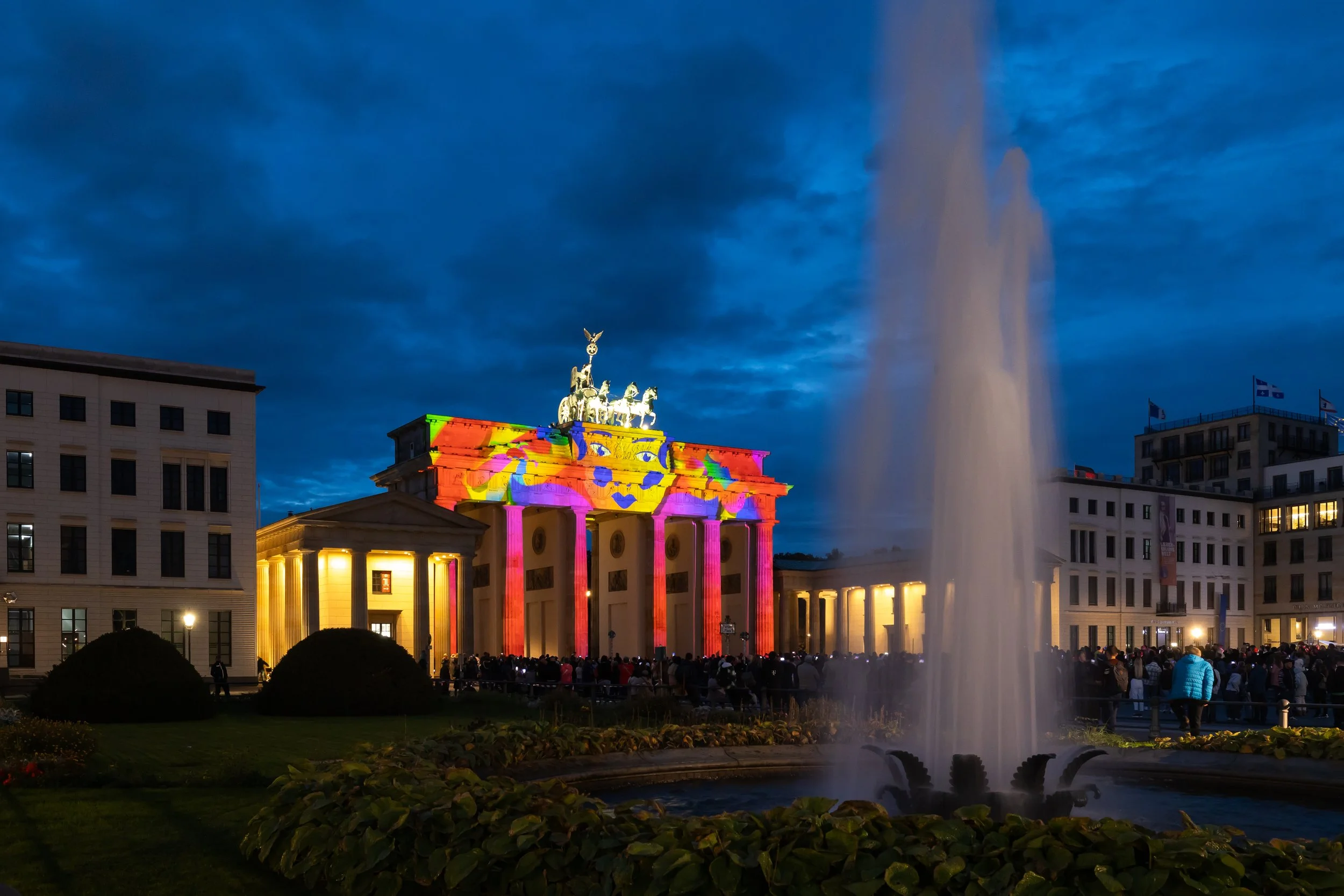 Nighttime scene of the Brandenburg Gate in Berlin illuminated with colorful lights projected onto it, with a fountain in the foreground and a crowd of people gathered nearby.