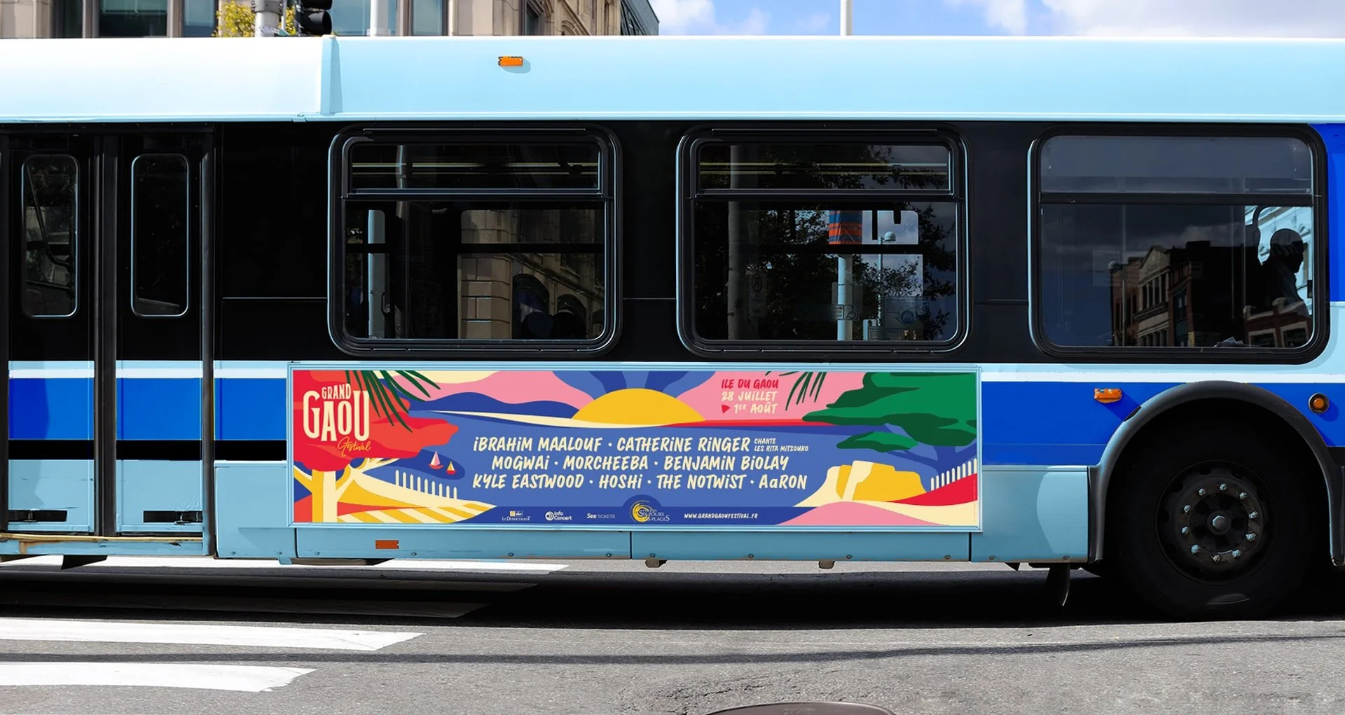 Blue bus with a colorful advertisement for the Grand Gaou festival, featuring names of performers and festival dates in July and August, in front of a city street.