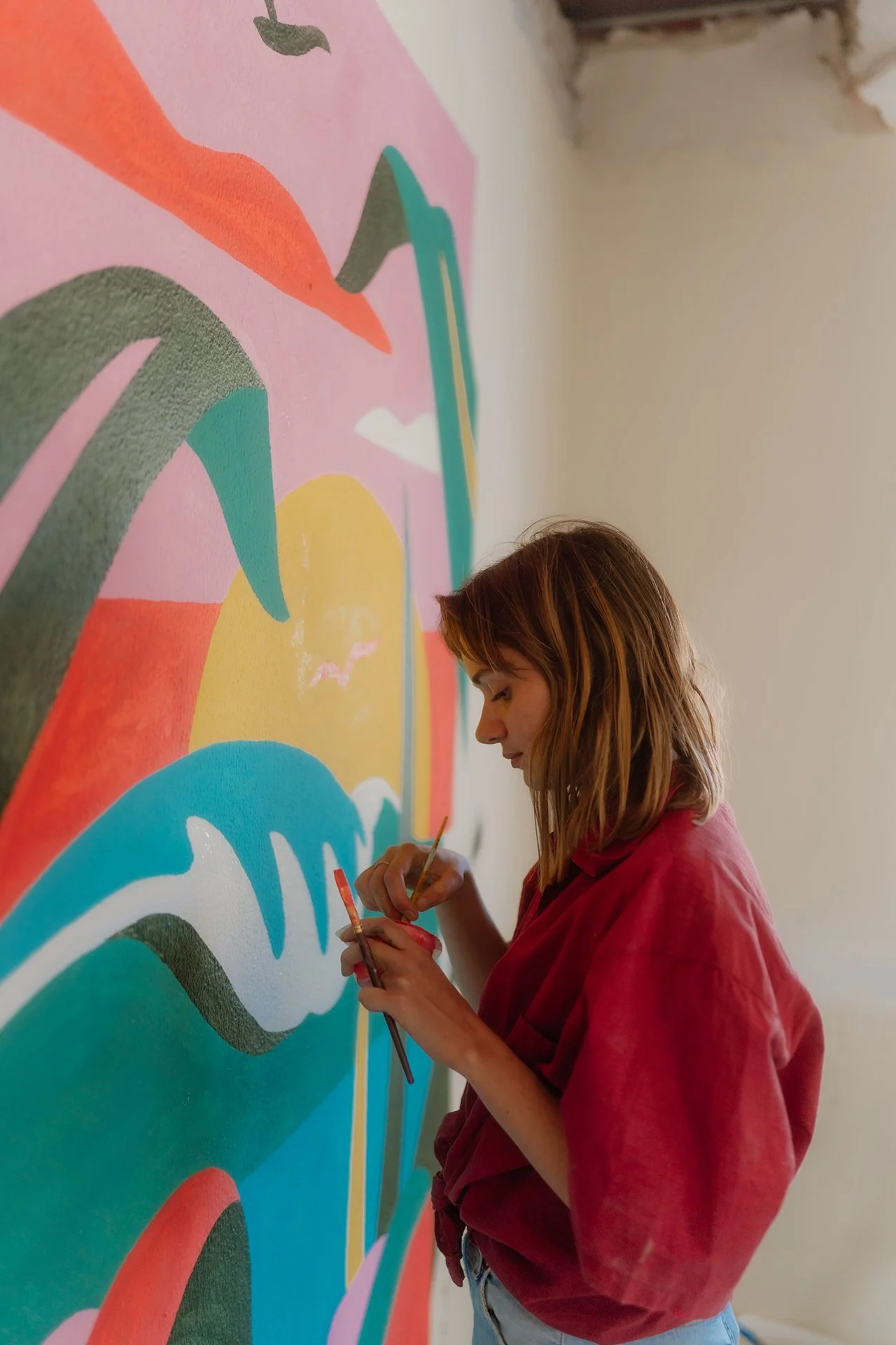 A woman in a red shirt is painting a colorful abstract mural on a wall.