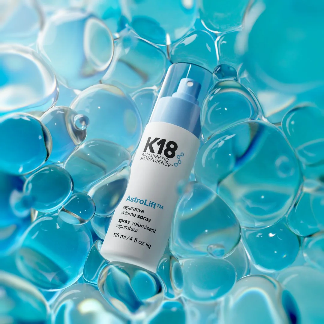 Ad artwork showcasing a white spray bottle labeled K18 Biomimetic Hairscience AstroLift Reparative Volume Spray surrounded by blue gel-like bubbles.
