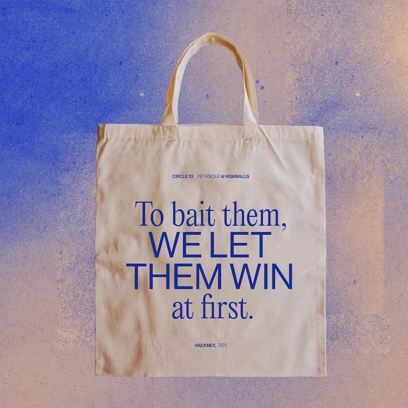 A beige tote bag with blue text printed on it, placed on a gradient background from blue to pink. The text reads 'To bait them, WE LET THEM WIN at first.'