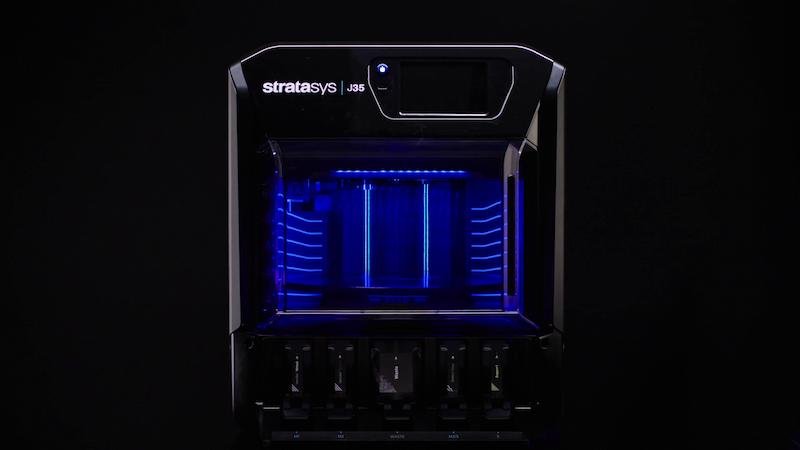 A large, black 3D printer with blue LED lighting, labeled 'stratasys J35', is shown against a dark background. Video shoot production done by Savage Films