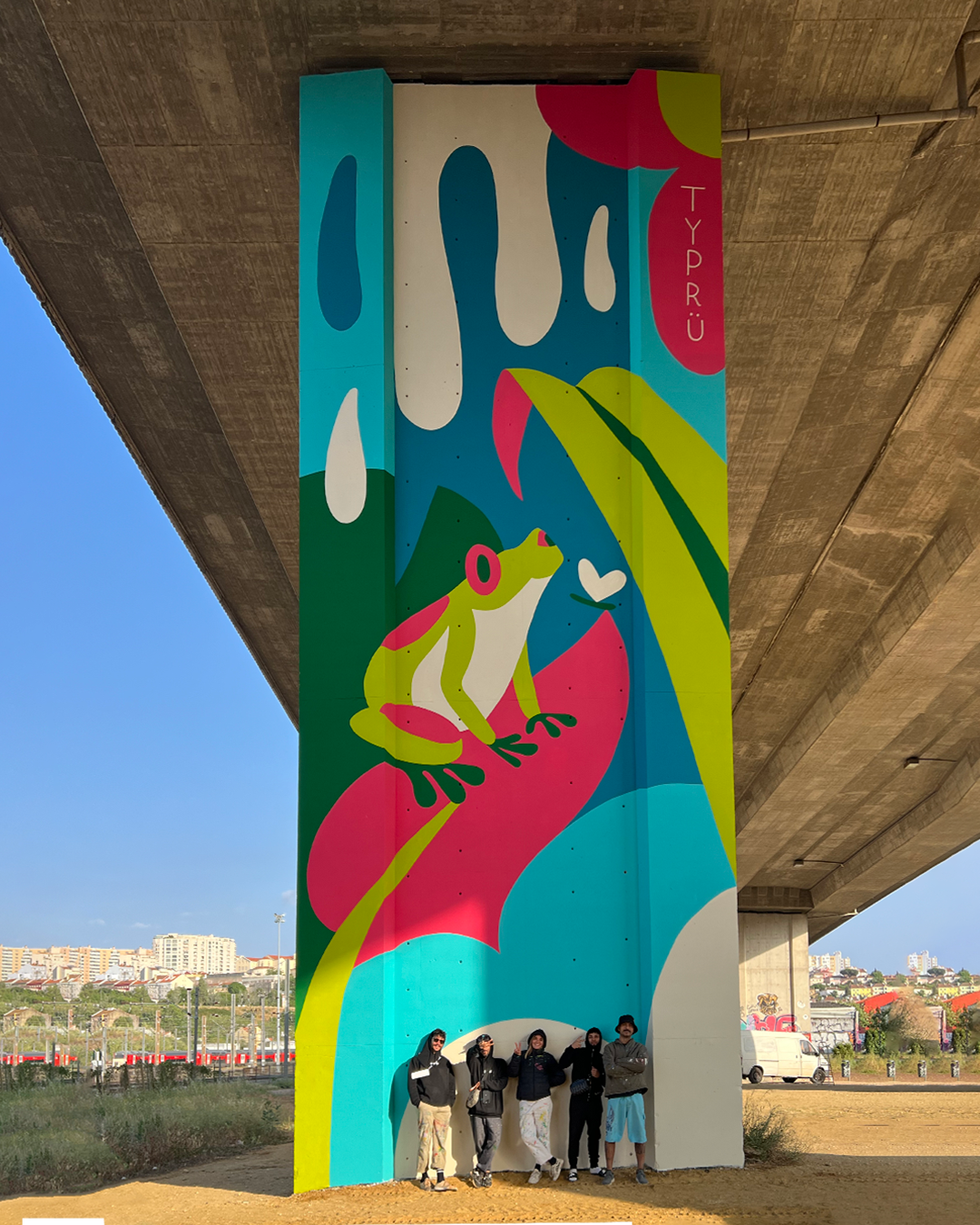 Brightly colored mural on a large pillar under a bridge, featuring a frog sitting on a pink leaf, surrounded by abstract shapes and hearts, with the word 'TYP.RÜ' written along the edge. A group of five people stands at the base of the pillar.