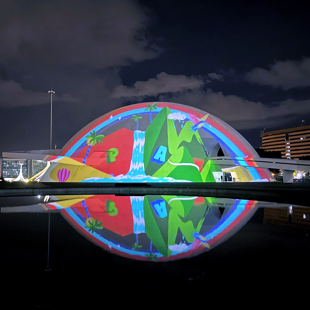 Nighttime view of a colorful, illuminated, dome-shaped building with a landscape scene projected onto it, including tropical trees, mountains, waterfalls, and clouds, with reflections visible in a pool of water in the foreground.
