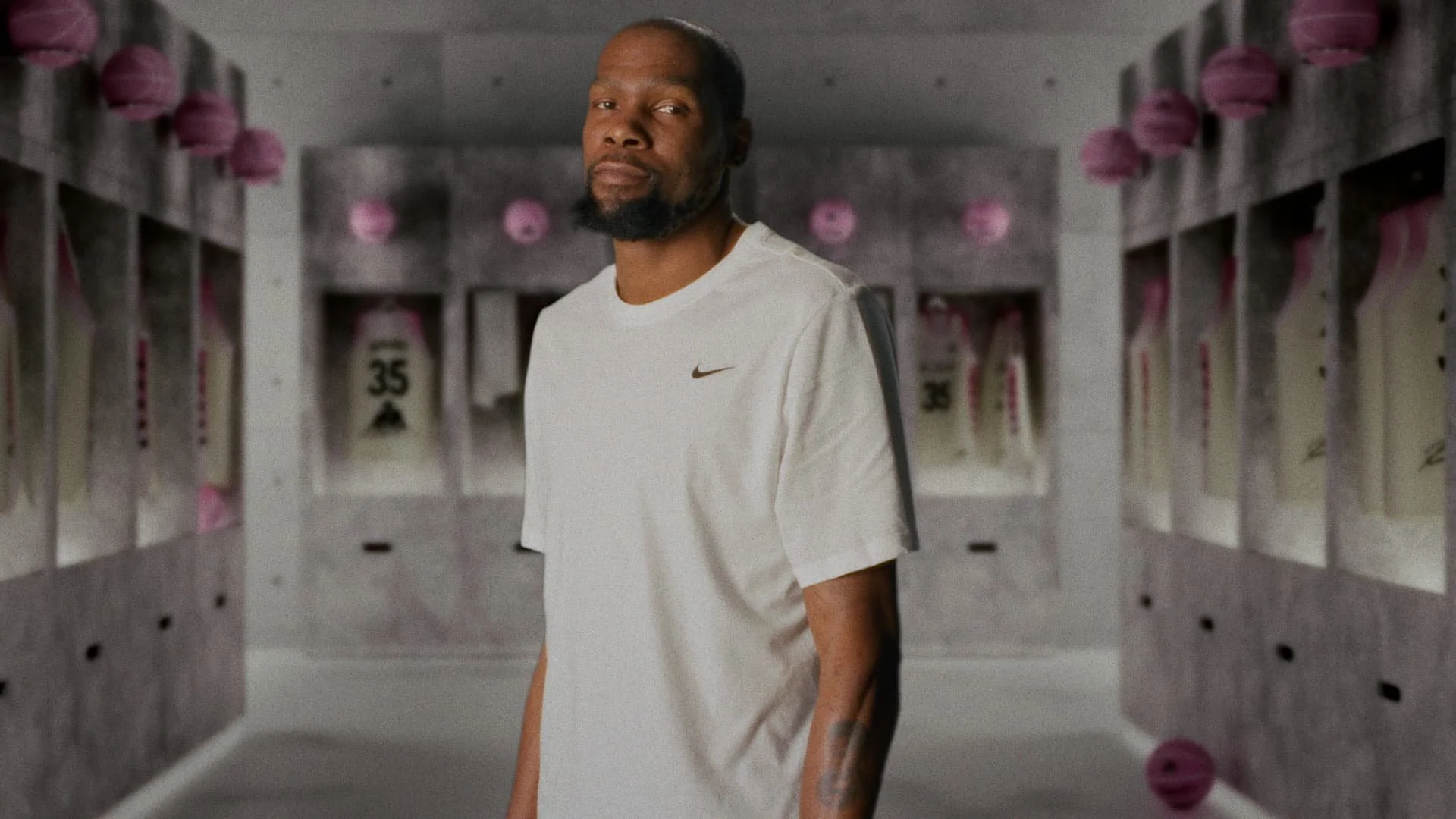 Man in a white Nike shirt standing in a locker room with jerseys displayed in the background.