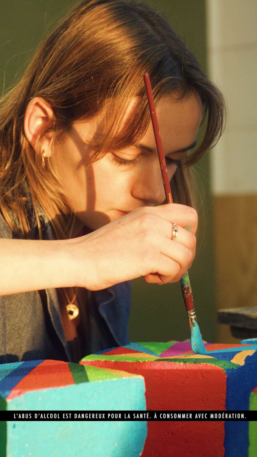 A young woman carefully paints the surface of a colorful, geometric-patterned object with a small brush.