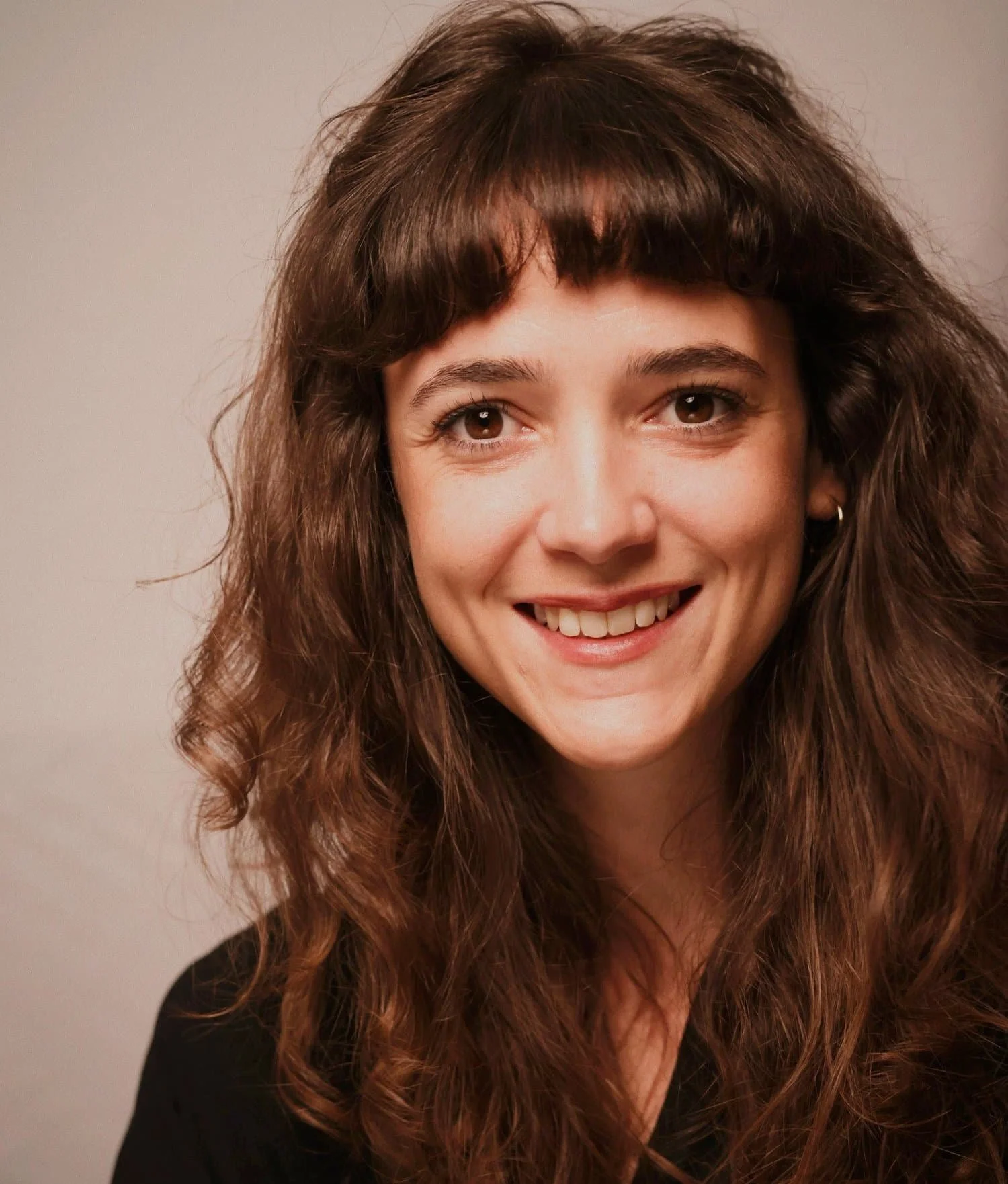 Portrait of a woman facing camera and smiling, with a fringe and long brown weavy hair