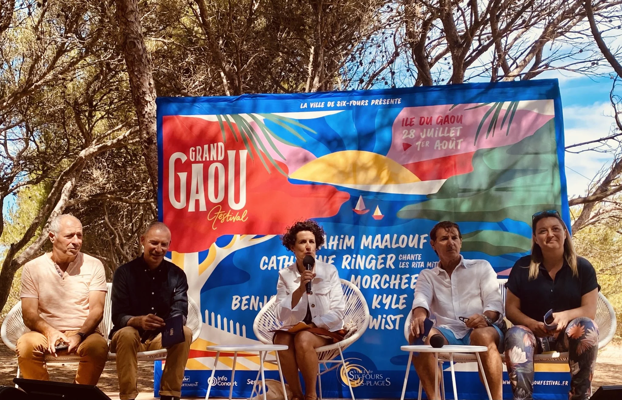 Panel of five people seated in front of a colorful banner for the Grand Gauf Festival, outdoors with trees in the background. The banner features a sunset scene and event details in French.