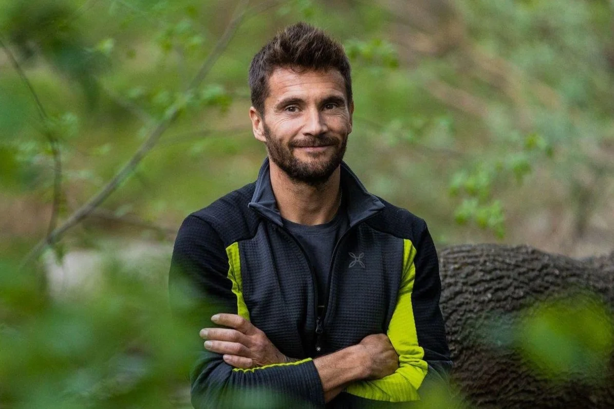 Man in outdoor clothing standing in a forest with arms crossed, smiling.