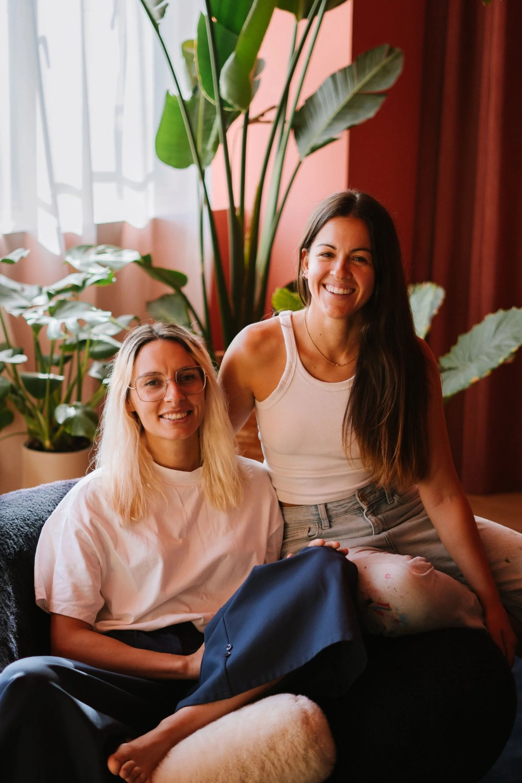 Two women sitting together indoors surrounded by plants, smiling, with one wearing glasses and a white shirt, and the other in a tank top and jeans.