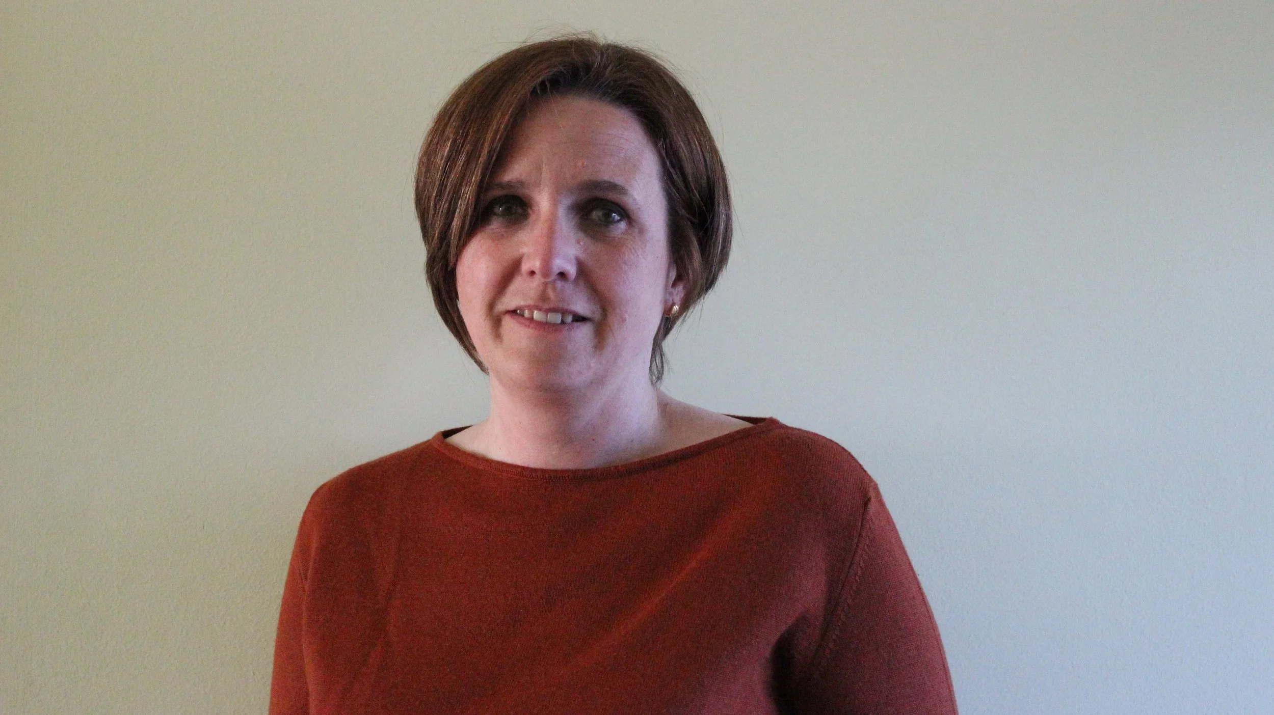 Person with short brown hair and a red sweater standing against a neutral background.