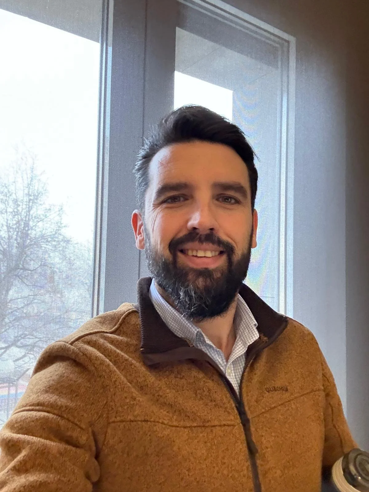 Man in brown sweater standing indoors in front of a window, smiling.