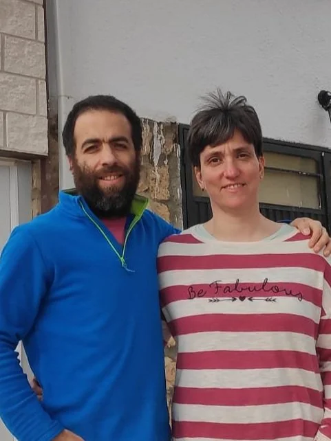 Two people standing close together, smiling, in front of a building. One is wearing a blue jacket, and the other is wearing a red and white striped shirt with the words "Be Fabulous."