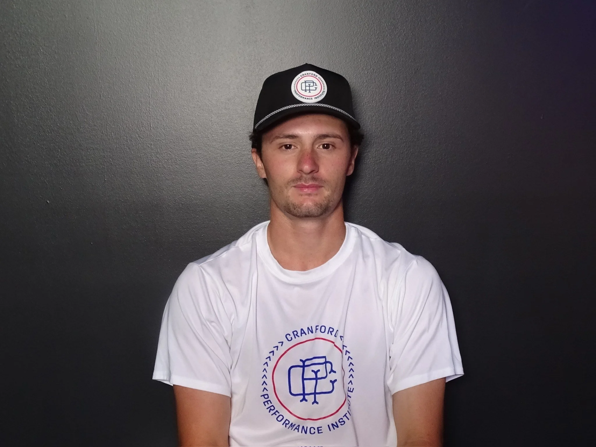 Young man with short dark hair, wearing a black cap with an embroidered logo, a white T-shirt with a circular logo reading 'Cranford Performance Institute' and a graphic of a gym, sitting against a dark gray wall.