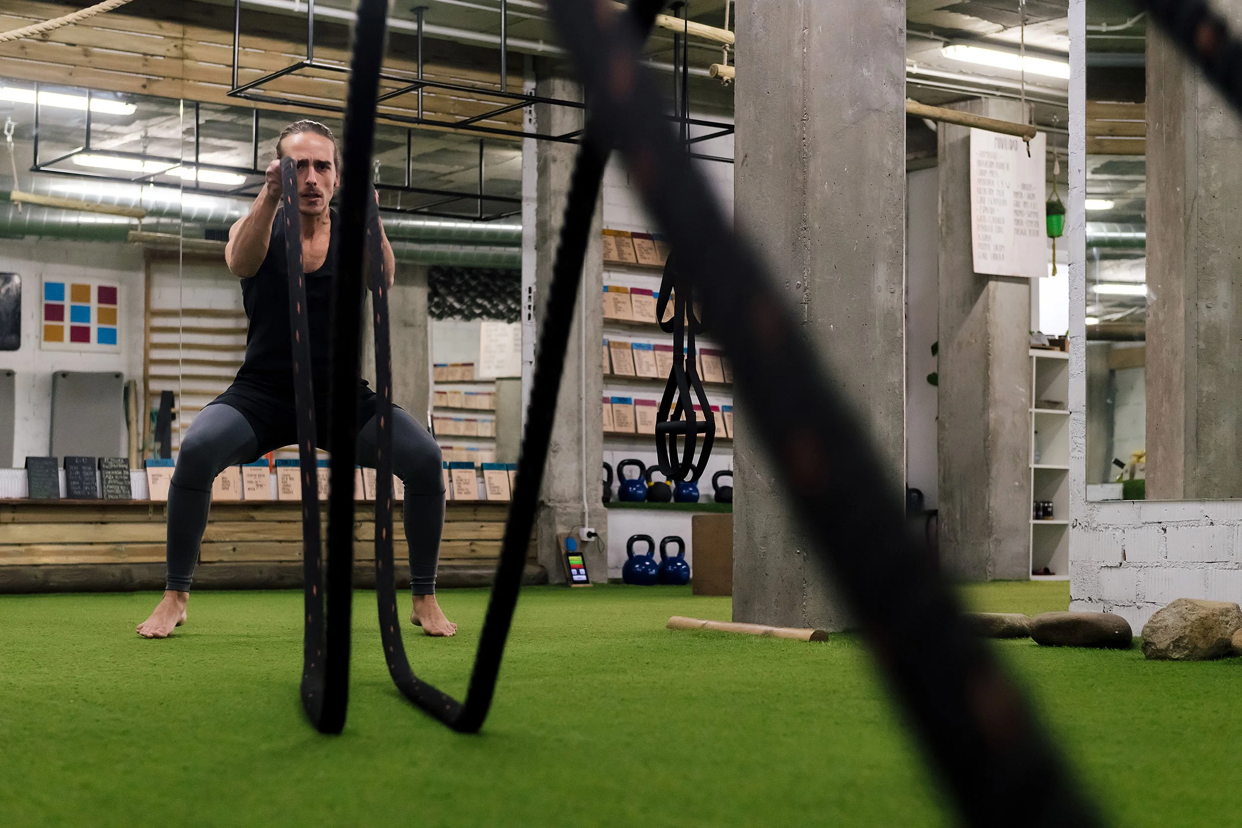 A woman working out in a gym, holding battle ropes with a focused expression on her face, on a green turf floor, with kettlebells and fitness equipment in the background.