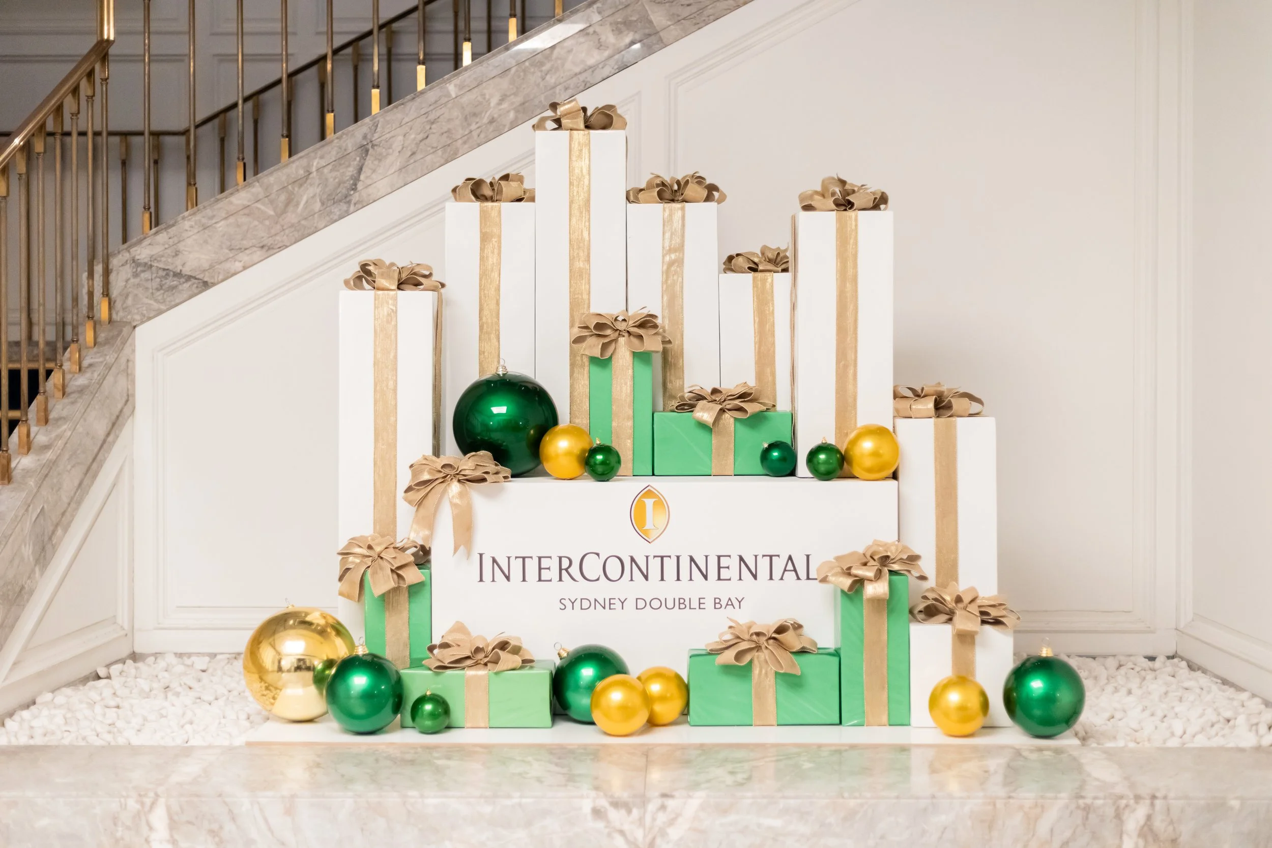 An artistic holiday installation featuring clean, geometric gift boxes in crisp white and deep emerald green stacked dramatically below a grand marble staircase.