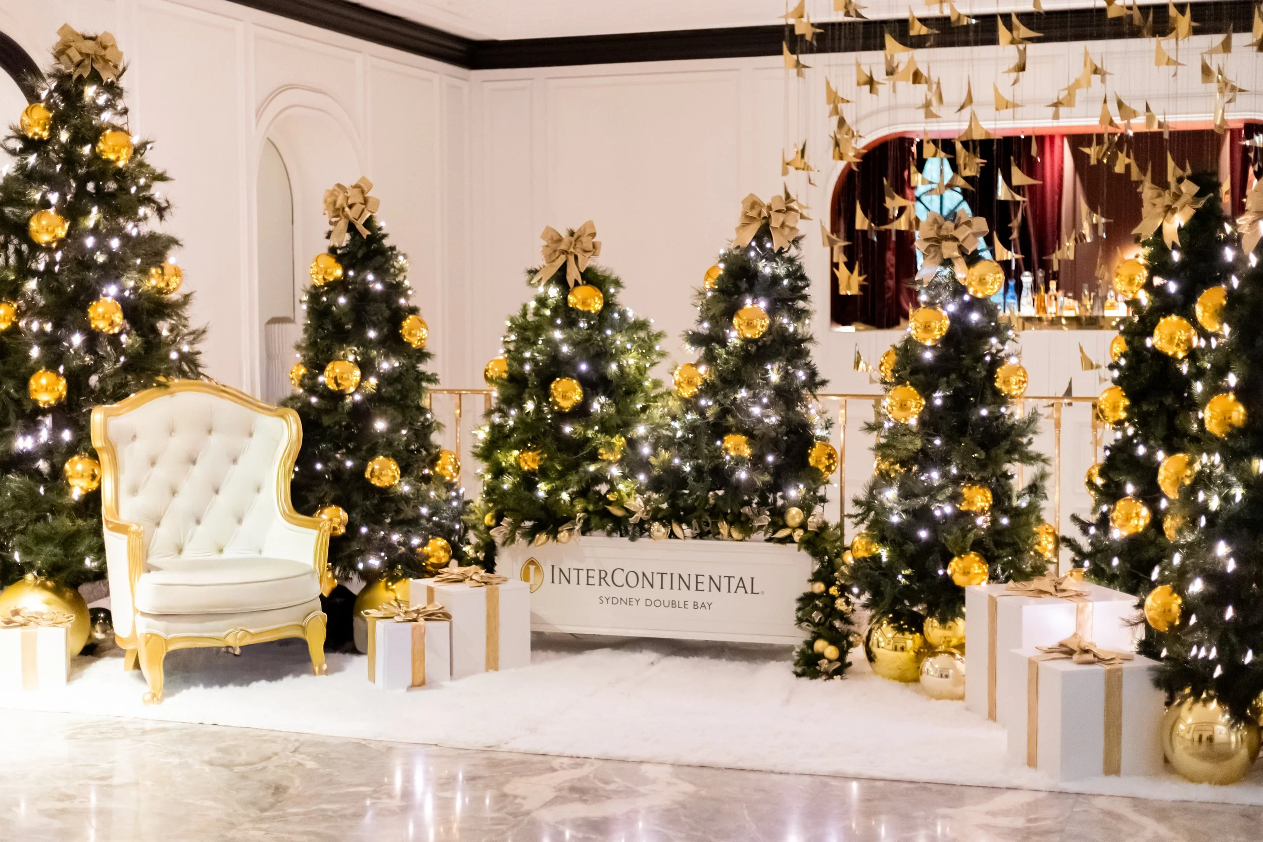 A close-up shot of expertly styled holiday trees capturing the soft, ambient glow of interwoven fairy lights and oversized reflective gold baubles.
