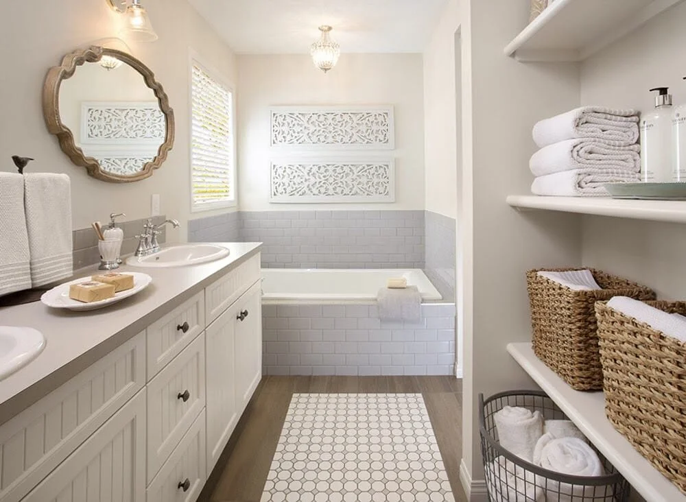 Modern bathroom with double vanity, two sinks, decorative mirror, white towels, wicker baskets, gray tiles around bathtub, and geometric rug.