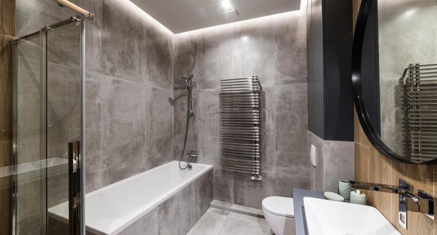 Modern bathroom with gray tiled walls, a white bathtub with a showerhead, a glass shower door, a wall-mounted towel heater, a toilet, a sink with a mirror, and ambient lighting.
