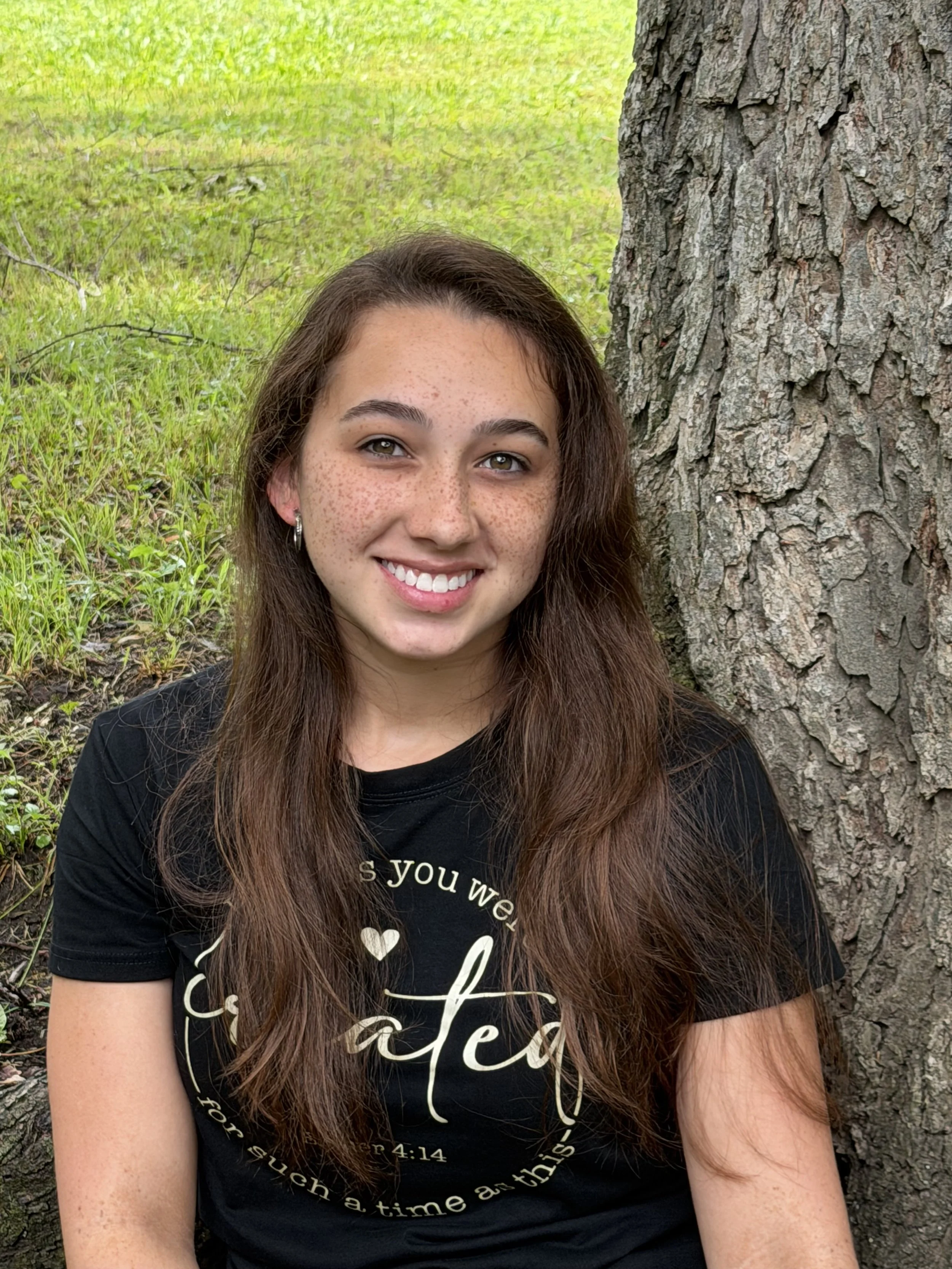 A young woman with long brown hair, freckles, and hoop earrings is sitting outdoors next to a large tree trunk, smiling at the camera. She is wearing a black T-shirt with white text and a heart design.