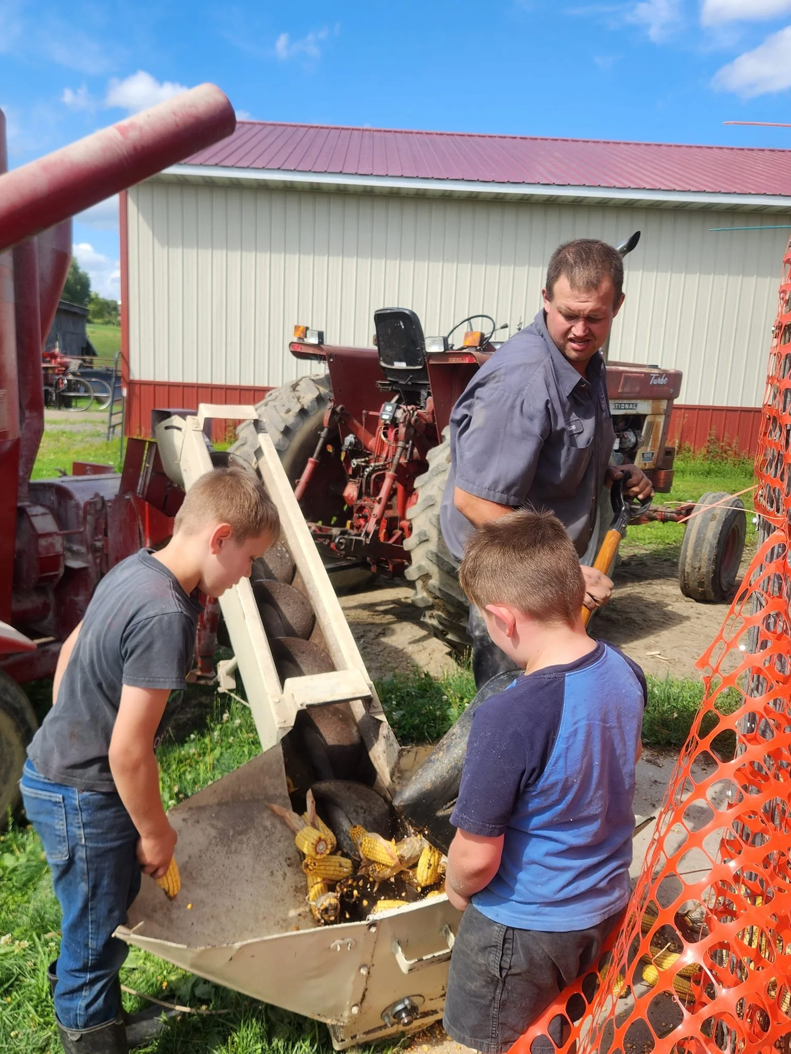 Three children and a man working with farm machinery outdoors, with a barn in the background, in bright weather.