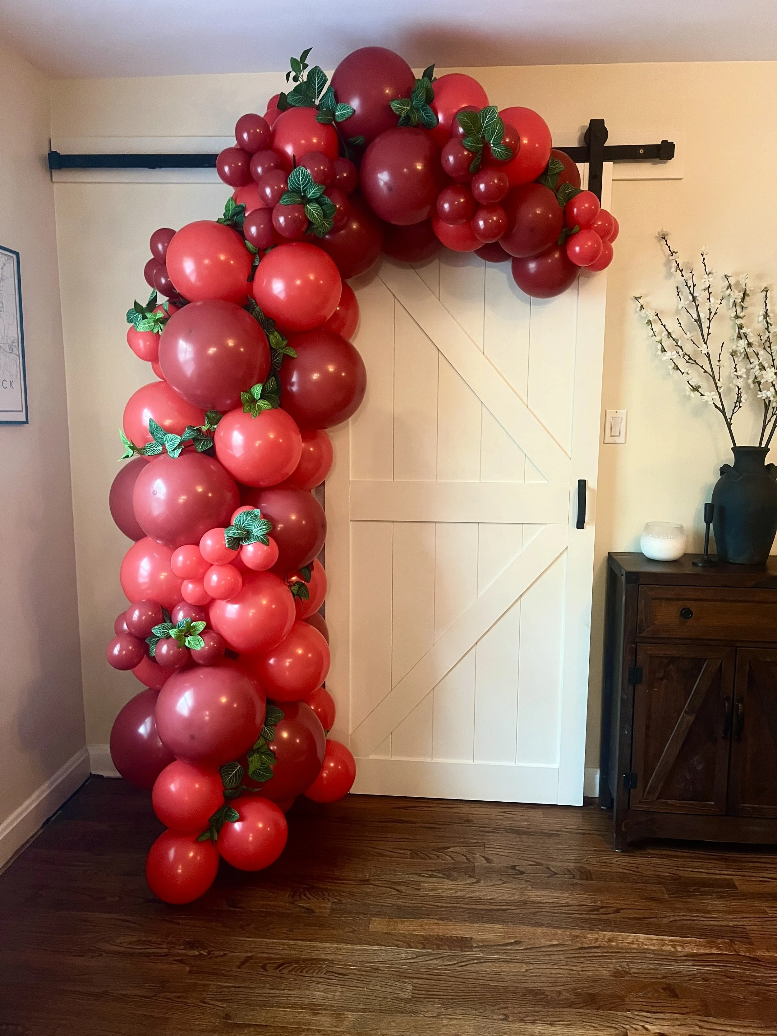 Decorative balloon arrangement with red and dark red balloons and green leaves, positioned near a white sliding barn door. Interior room with hardwood flooring, a dark wood cabinet, and a vase with white flowering branches.