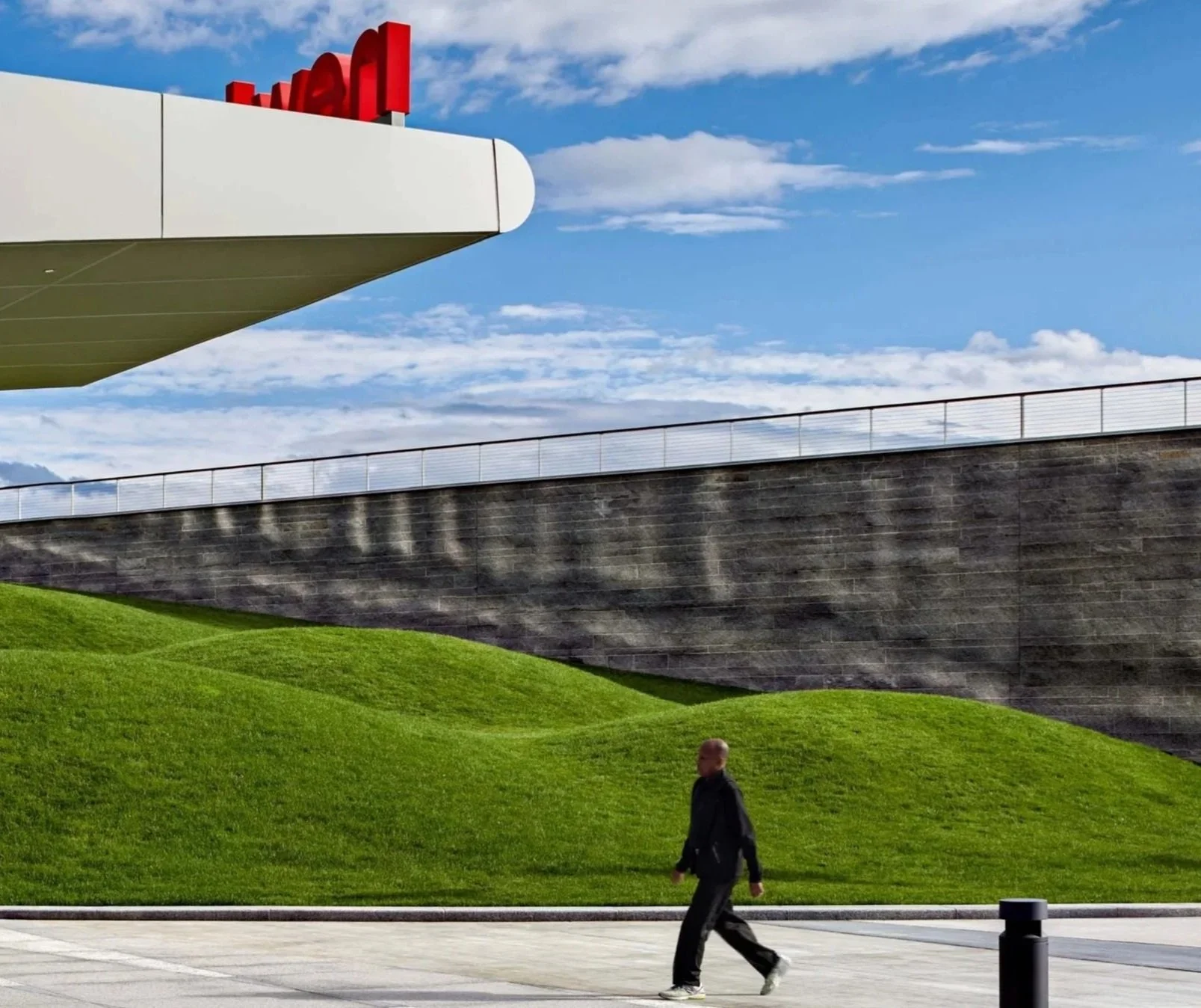 Person walking past a modern building with red rooftop lettering, a stone retaining wall, and grassy landscaping under a blue sky